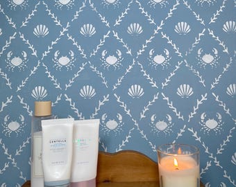 May include: A wooden shelf holds skincare products and a lit candle in a glass. The background is a blue wall with a white crab and seashell pattern. The products include a clear bottle and two tubes with white labels.