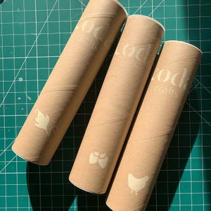Puede incluir: Tres tubos de env&iacute;o cil&iacute;ndricos de cart&oacute;n en color marr&oacute;n claro. Cada tubo presenta el texto "coda crafts" impreso en color crema cerca de la parte superior, junto con un icono gr&aacute;fico &uacute;nico: una hoja, dos fresas y un pollo.