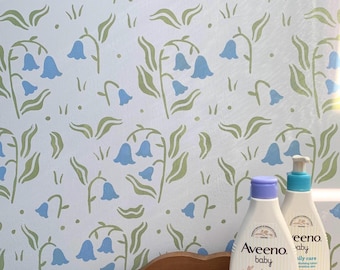 May include: Two Aveeno baby calming comfort bottles with blue and purple caps sit on a wooden shelf. The background features a white wall with a bluebell floral pattern in blue and green.