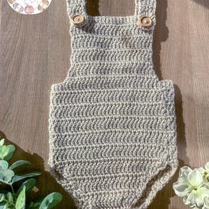 May include: A crocheted baby romper in a beige color with wooden buttons. The romper has a chevron pattern and is made with a soft yarn.
