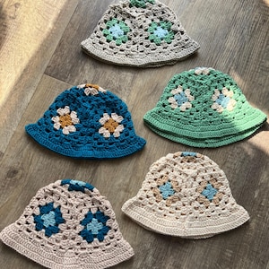Könnte beinhalten: Fünf gehäkelte Fischerhüte in verschiedenen Farben: Blau, Grün, Beige und Grau. Jeder Hut hat ein Blumenmuster in Kontrastfarben. Die Hüte sind auf einer Holzoberfläche angeordnet und zeigen ihre handgefertigte Qualität.