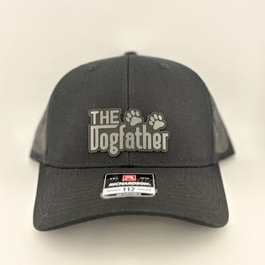 May include: Black trucker hat with the text "The Dogfather" in gray lettering and paw prints. The hat has a curved brim and a mesh back. The Richardson 112 hat is adjustable.