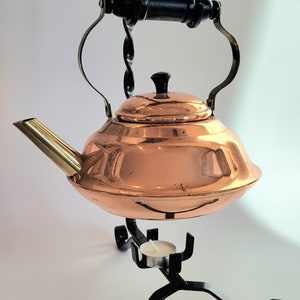 May include: A copper teapot with a black handle and a brass spout sits on a black metal stand with a candle holder.