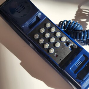 May include: A blue rotary phone with a black keypad and a coiled blue cord. The keypad has numbers 1 through 9, 0, *, and #. The phone has a small red light that says "in use".
