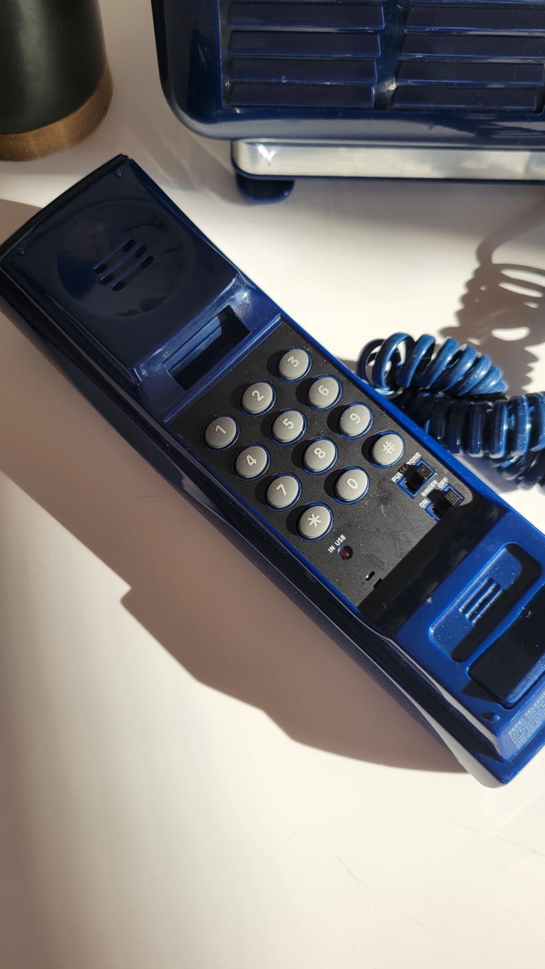 May include: A blue rotary phone with a black keypad and a coiled blue cord. The keypad has numbers 1 through 9, 0, *, and #. The phone has a small red light that says "in use".