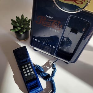 May include: A blue and yellow vintage-style Pepsi-Cola refrigerator with a blue rotary phone attached. The refrigerator has the text "Drink Pepsi-Cola" and "Ice Cold" on it.
