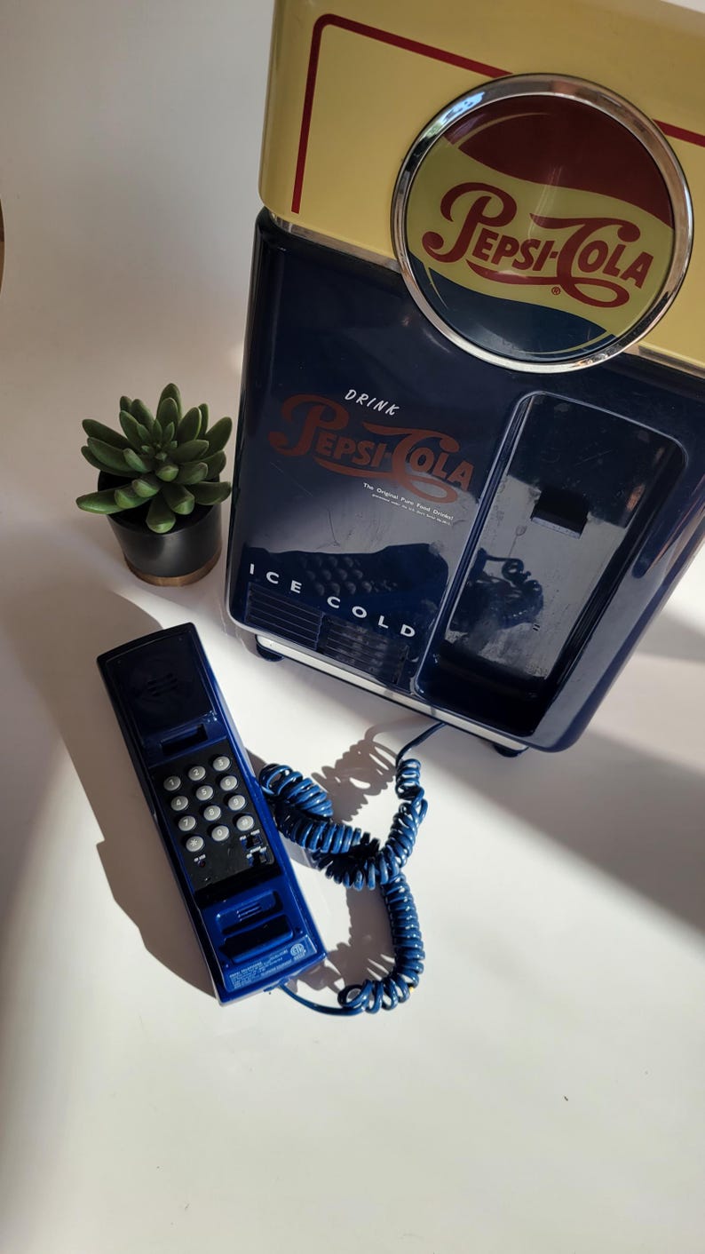May include: A blue and yellow vintage-style Pepsi-Cola refrigerator with a blue rotary phone attached. The refrigerator has the text "Drink Pepsi-Cola" and "Ice Cold" on it.