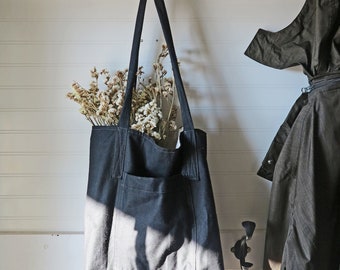 May include: A black canvas tote bag with a front pocket hangs from a black hook on a white paneled wall. The bag is filled with dried flowers. A dark coat hangs nearby.