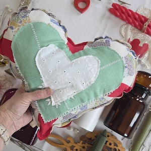May include: A handmade heart-shaped pillow with a patchwork design. The pillow features a green center heart with a white embroidered heart on top. The edges are trimmed with red fabric and a ruffled border. The pillow is held in a person's hand.