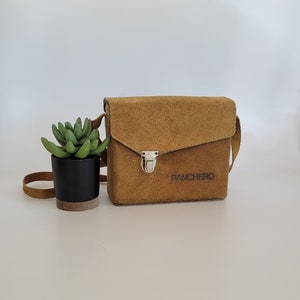 May include: A brown suede camera bag with a silver clasp and the word "Ranchero" printed on the front.
