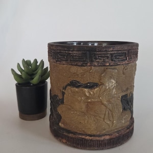 May include: A cylindrical, decorative planter with a relief design. The planter is primarily tan with dark brown accents and a textured surface. A small succulent in a black pot sits to the left.