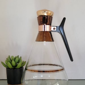 May include: A vintage glass carafe with a conical shape, featuring a gold-colored lid and trim. The carafe has a black handle and a clear body, with a gold band around the middle. A small green plant in a black pot sits to the left.