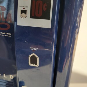 May include: A blue vintage soda vending machine with a coin return slot, a 10 cent price tag, and a drop bottle cap here slot. The machine has a white label that says "Cola Food Drinks!"