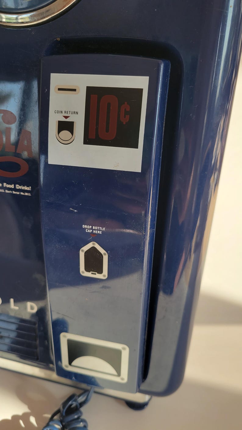 May include: A blue vintage soda vending machine with a coin return slot, a 10 cent price tag, and a drop bottle cap here slot. The machine has a white label that says "Cola Food Drinks!"