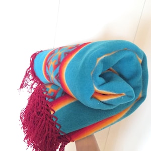 May include: A rolled blanket featuring a vibrant turquoise base color with horizontal bands of orange and red. The blanket displays a geometric pattern in red and orange along the edges and is finished with long, deep red fringe tassels.