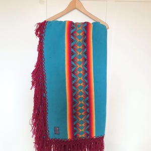 May include: A turquoise blanket with a central vertical stripe in red, orange, and yellow geometric patterns. The blanket has a burgundy fringe along the edges and is hanging on a wooden hanger.