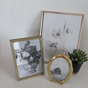 May include: Three gold-toned picture frames of varying sizes, each holding a black and white image. One frame is rectangular, another is square, and the third is oval. A small potted succulent sits nearby.