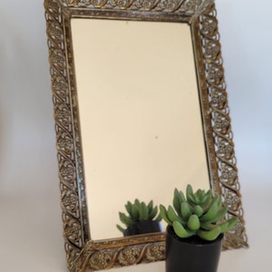 May include: A gold-toned metal tray with a decorative, ornate frame and a mirror surface. A small potted succulent plant sits in front of the tray.