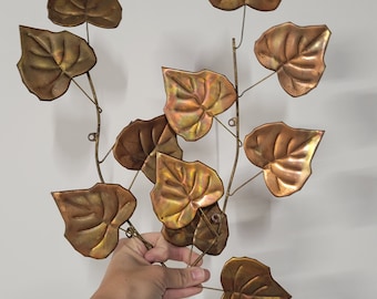 May include: A wall hanging sculpture made of copper leaves, with a hand holding the sculpture in front of a white wall.