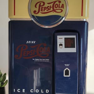 May include: A blue and yellow vintage-style Pepsi-Cola phone with a red and blue Pepsi logo on the top. The phone has a coin return slot and a drop bottle cap here slot. The phone says "Drink Pepsi-Cola The Original Pure Food Drink!" and "Ice Cold".