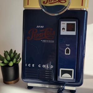 May include: A blue and yellow vintage-style Pepsi-Cola phone with a rotary dial and a cord. The phone has a sign that says "Drink Pepsi-Cola, The Original Pure Food Drink!" and "Ice Cold".