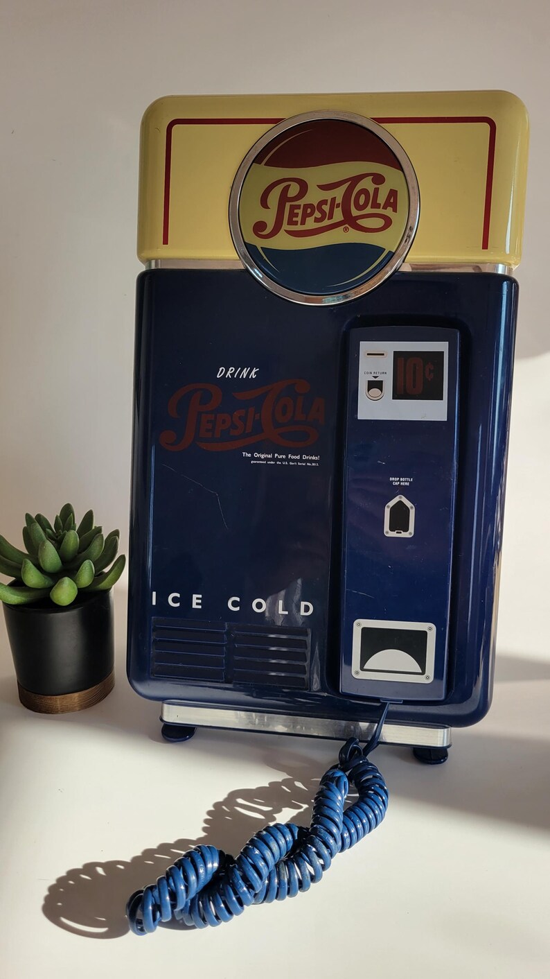 May include: A blue and yellow vintage-style Pepsi-Cola phone with a rotary dial and a cord. The phone has a sign that says "Drink Pepsi-Cola, The Original Pure Food Drink!" and "Ice Cold".