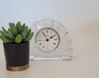 May include: A clear crystal clock with Roman numeral markings and a white face. The clock is on a white surface next to a small potted succulent plant.