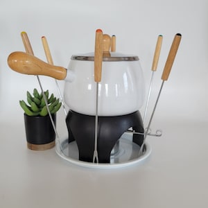 May include: White ceramic fondue pot with a black stand and a white plate. The pot has a wooden handle and six wooden fondue forks with colored tips.