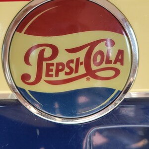 May include: A vintage Pepsi-Cola vending machine with a red, white, and blue logo. The machine is painted blue and has a yellow top with a red stripe. The text "DRINK PEPSI-COLA" is visible on the front of the machine.