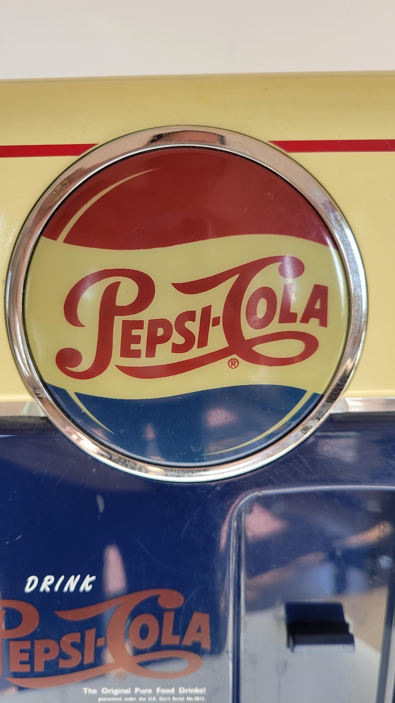May include: A vintage Pepsi-Cola vending machine with a red, white, and blue logo. The machine is painted blue and has a yellow top with a red stripe. The text "DRINK PEPSI-COLA" is visible on the front of the machine.