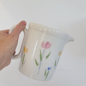 May include: White ceramic pitcher with a handle and a floral design. The flowers are painted in shades of pink, yellow, blue, and green.