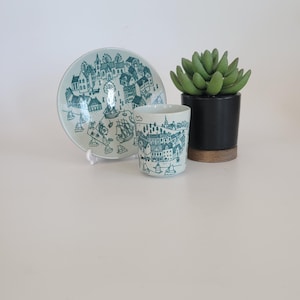 May include: A ceramic plate and cup set with a teal townscape design. The plate is round and supported by a white stand. The cup is cylindrical. A small succulent plant in a black pot with a wooden base is also present.