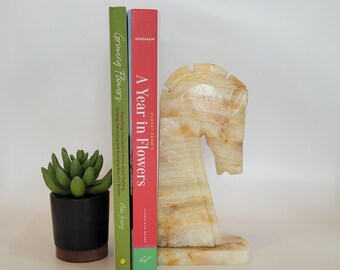May include: A pair of bookends shaped like a horse's head, made of white and yellow marble. The bookends are holding up two books, one with a green cover and the other with a red cover. The book with the green cover has the title "Growing Flowers" and the book with the red cover has the title "A Year in Flowers".
