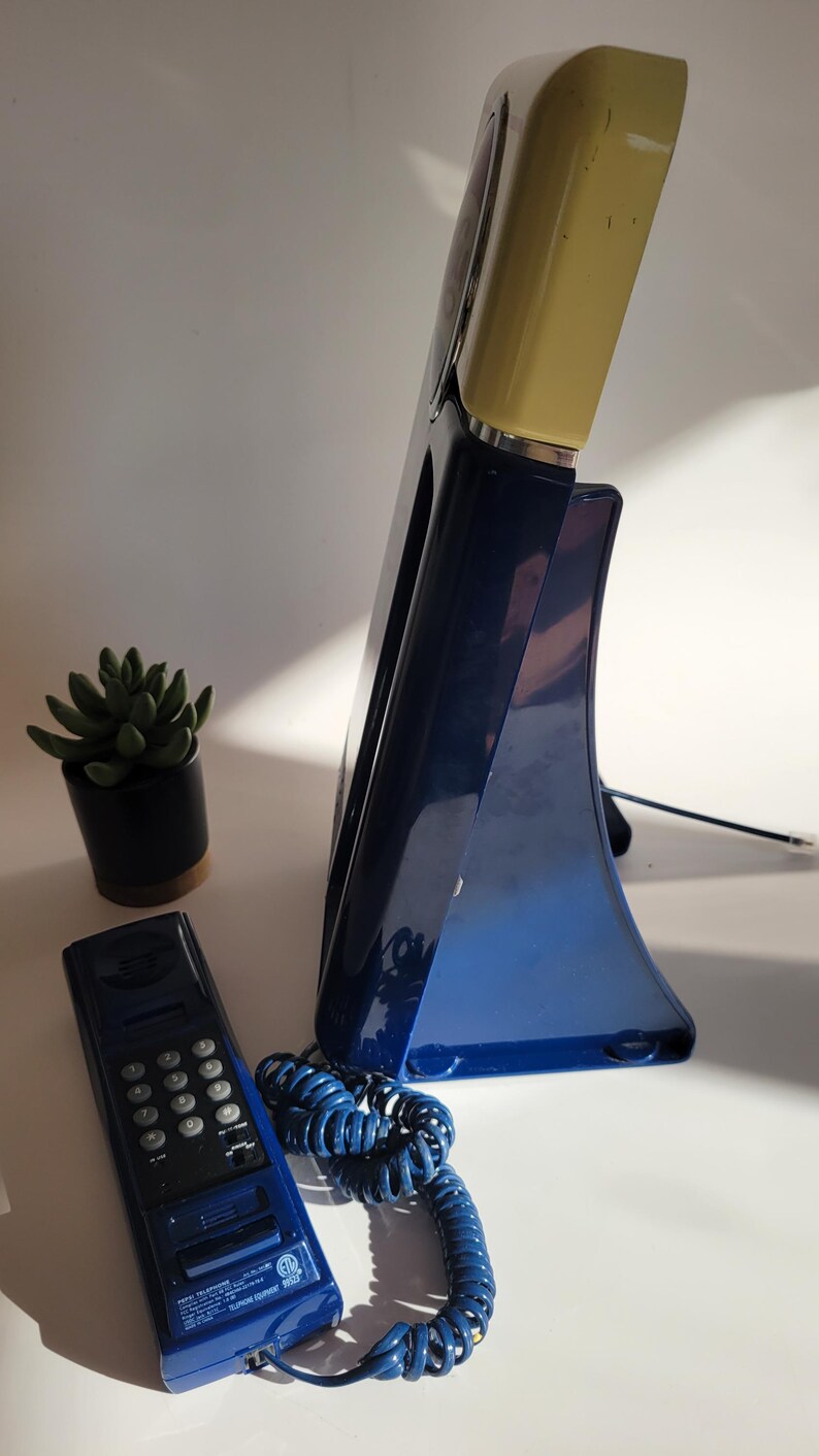 May include: A blue rotary phone with a yellow base and a separate handset. The phone has a coiled cord and a keypad on the handset.