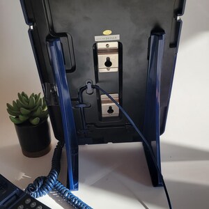 May include: A blue corded phone with a rotary dial and a separate base unit. The base unit has a blue stand and a black back panel with a serial number label. The phone is connected to the base unit with a coiled cord.