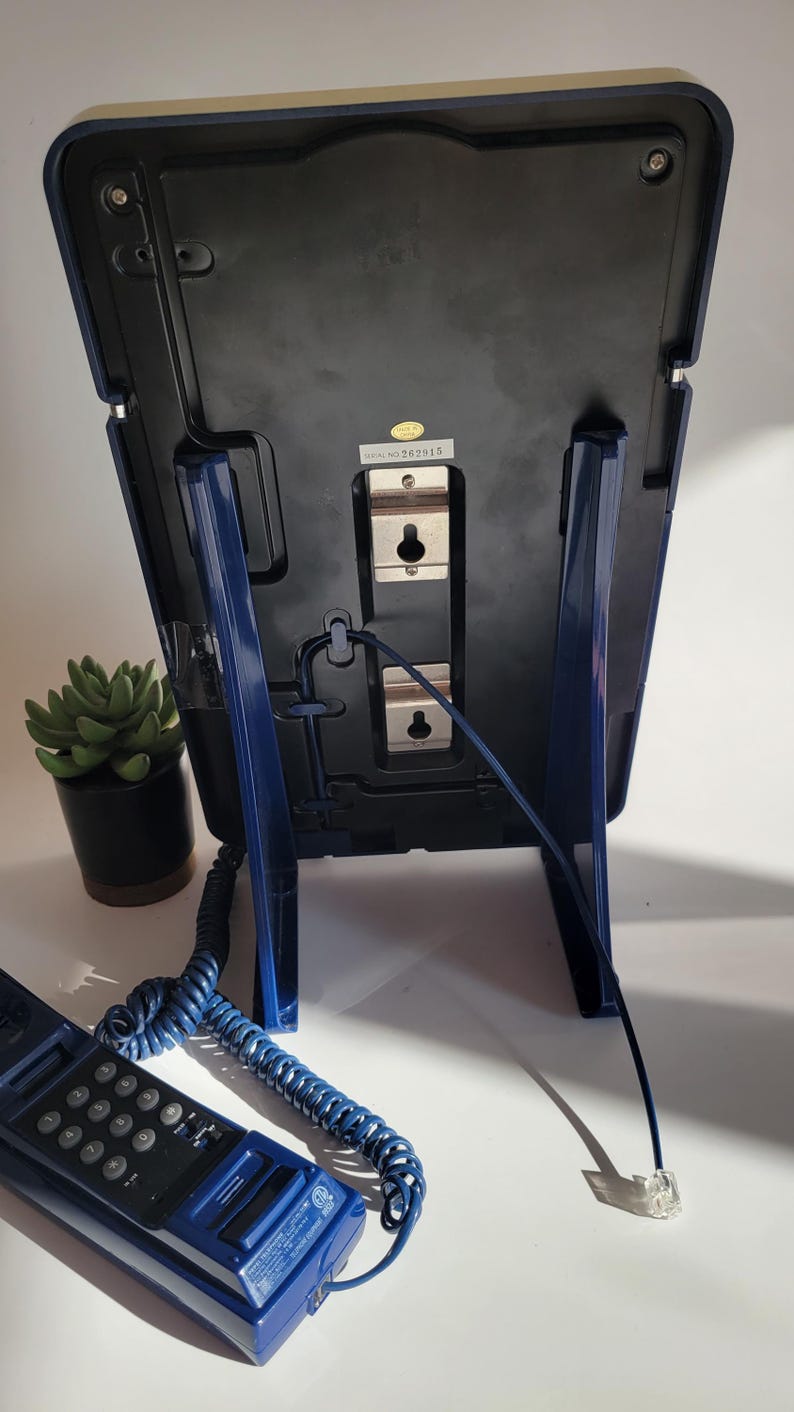 May include: A blue corded phone with a rotary dial and a separate base unit. The base unit has a blue stand and a black back panel with a serial number label. The phone is connected to the base unit with a coiled cord.