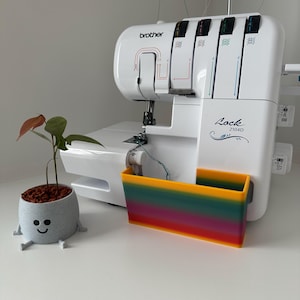 Könnte beinhalten: Eine weiße Brother Lock 2104D Overlock-Nähmaschine mit einem regenbogenfarbenen Abfallbehälter. Links ein kleiner grauer Pflanztopf mit einem lächelnden Gesicht und einer Pflanze mit grünen und roten Blättern.