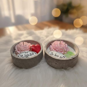 Peut inclure: Deux bougeoirs en béton avec des motifs floraux. L'un a une bougie rose en forme de rose et un cœur rouge, avec le texte "Ich liebe Dich". L'autre a une bougie rose avec le texte "Du bist toll".
