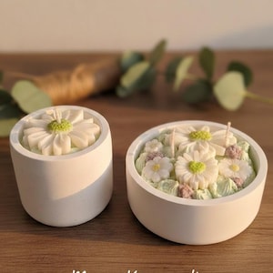 Könnte beinhalten: Zwei weiße, gänseblümchenförmige Kerzen in runden, weißen Behältern. Die Kerzen haben grüne und violette Akzente, und der Text "Mamas Kerzenwelt - Handmade Candles" steht unten. Die Kerzen stehen auf einer Holzoberfläche.