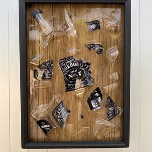 May include: A framed artwork featuring a wooden background with broken pieces of glass bottles and a Jack Daniel's label. The glass pieces are embedded in the wood and covered with resin.