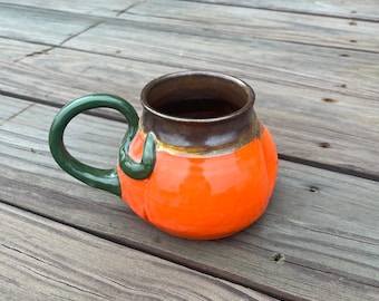 May include: Orange ceramic mug shaped like a pumpkin with a green handle. The mug has a brown rim and a glossy finish. The handle is a curved, solid green. The mug is sitting on a wooden surface.
