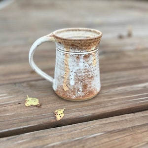 May include: A handmade ceramic mug with a white and brown speckled glaze. The mug has a large handle and a slightly flared rim. The exterior features a textured, rustic appearance. The mug is approximately 10 cm tall and is suitable for hot beverages.