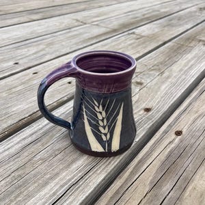 Puede incluir: Una taza de cerámica hecha a mano con una base azul oscuro y un borde y asa morados. La taza presenta un diseño de espiga de trigo en pintura color crema. La taza está sobre una superficie de madera.