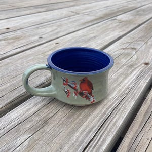 May include: A ceramic mug with a light green exterior, a blue interior, and a handle. The mug has a hand-painted design of a red cardinal bird on a branch. The mug is on a wooden surface. The mug's measurements are in centimetres.