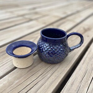 May include: A blue ceramic mug with a textured, iridescent glaze. The mug has a round body and a handle. A matching blue ceramic lid is also shown.