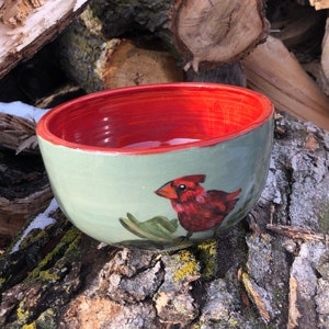 May include: A ceramic bowl with a red interior and a green exterior. The bowl features a painted cardinal bird with a red body and black eyes.
