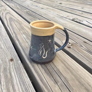 Puede incluir: Una taza de cerámica hecha a mano con un cuerpo azul oscuro y un borde beige. La taza presenta una silueta de gato blanca y diseños de estrellas. El asa es azul oscuro. La taza está sobre una superficie de madera.