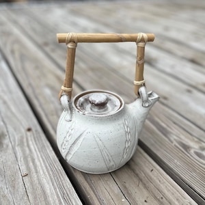 Puede incluir: Una tetera de cerámica blanca con asa de bambú. La tetera tiene un cuerpo redondo con tapa y boquilla. El asa está hecha de dos piezas de bambú unidas por hilo. La tetera tiene una superficie texturizada con diseños de espigas de trigo.
