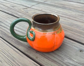 May include: A ceramic mug shaped like a pumpkin, with an orange body and a dark brown rim. The handle is green and has a curved, vine-like design. The mug is sitting on a wooden surface.