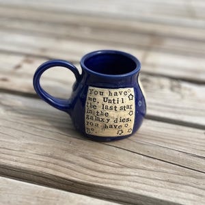May include: A small, dark blue ceramic mug with a rounded handle. The mug features a cream-colored rectangular patch with the text "You have me. Until the last star in the galaxy dies, you have me." and star illustrations.